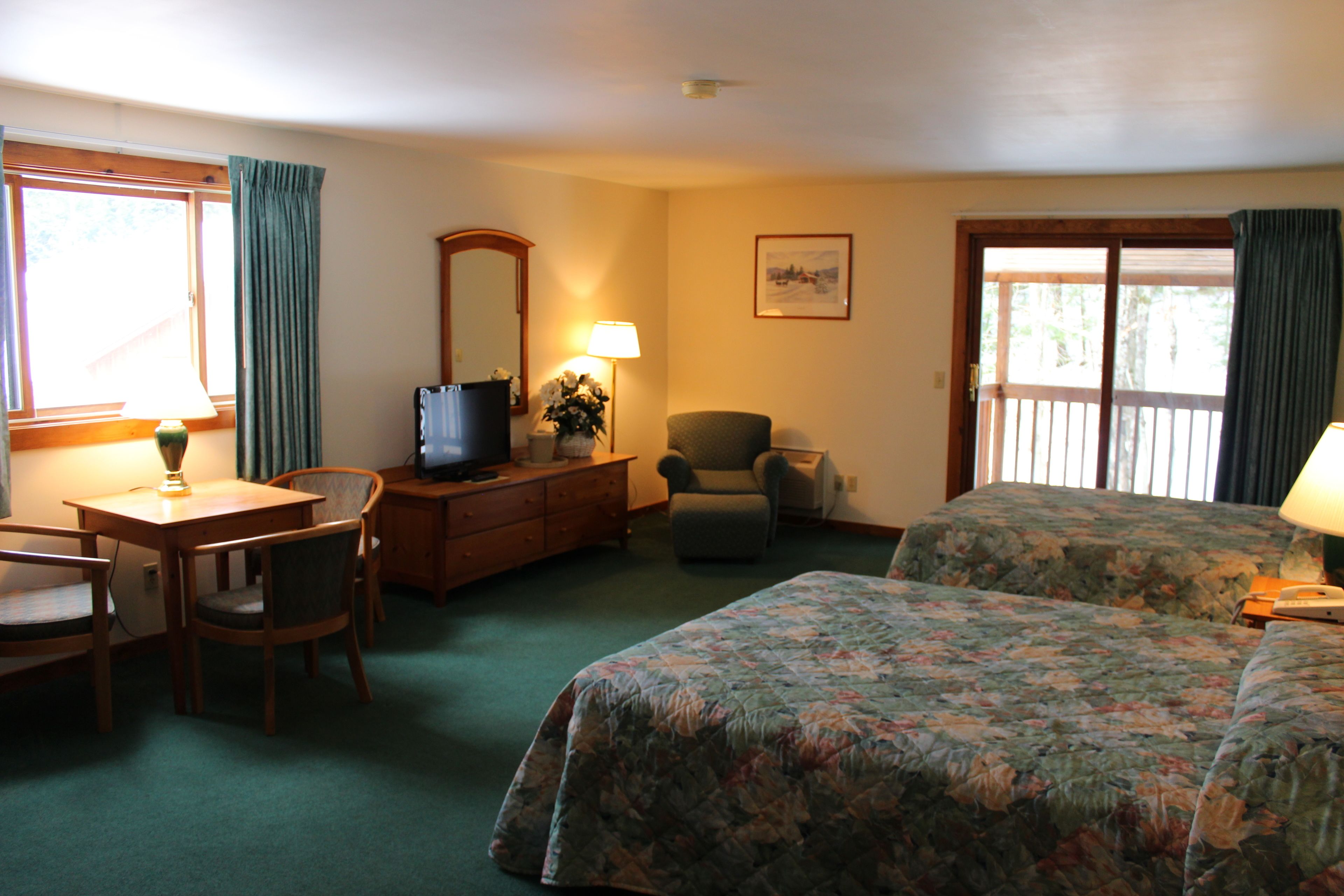 A hotel room with a desk, two chairs, a television, a dresser, a chair, a bed with floral bedding, a lamp, and a window with curtains.