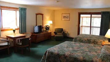 Quarto de hotel com escrivaninha, duas cadeiras, televisão, penteadeira, mais uma cadeira, cama com roupa de cama floral, abajur e janela com cortinas.