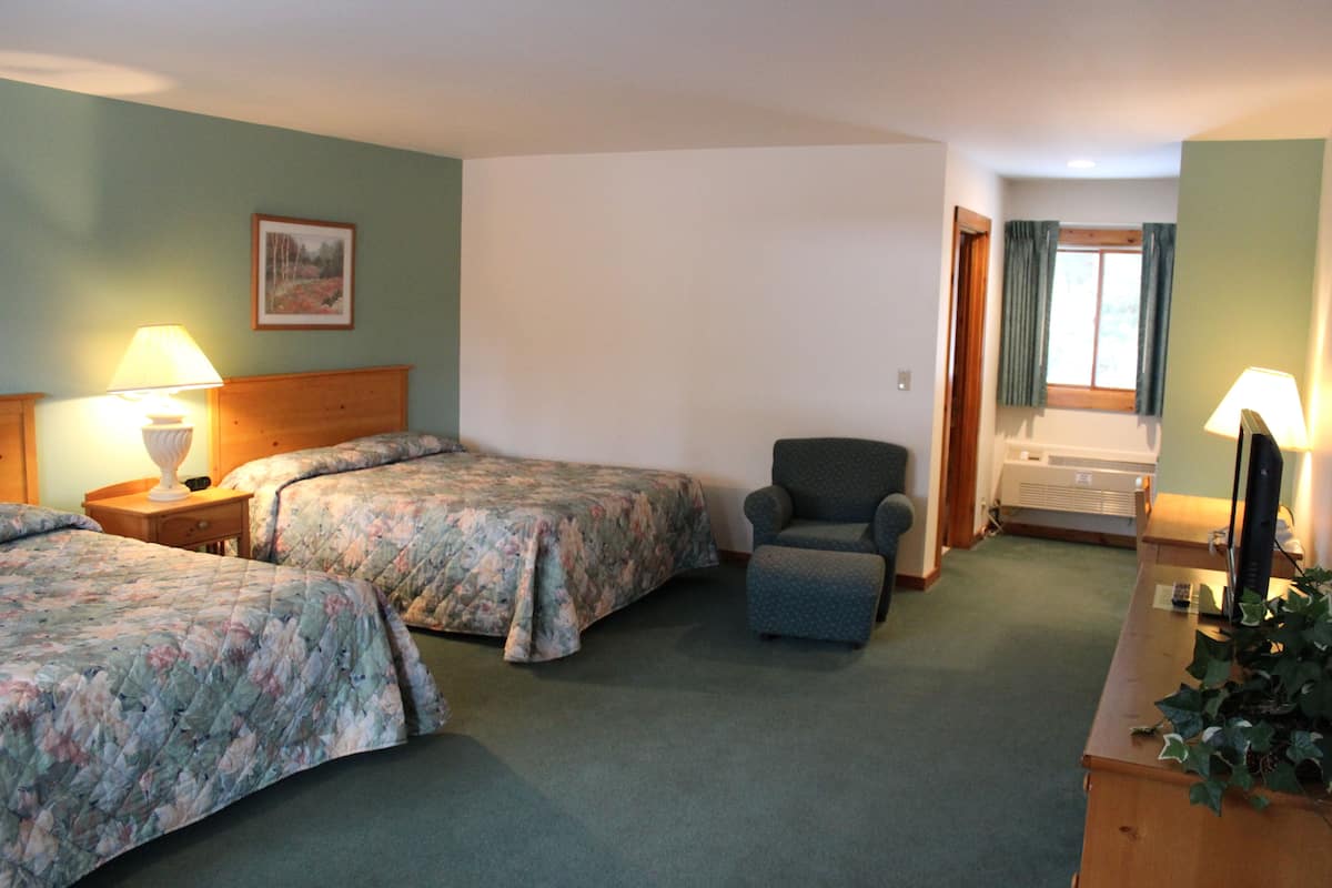 Ein Hotelzimmer mit zwei Betten, einem Sessel, einem Schreibtisch mit Lampe und einem fenster mit Vorhängen.
