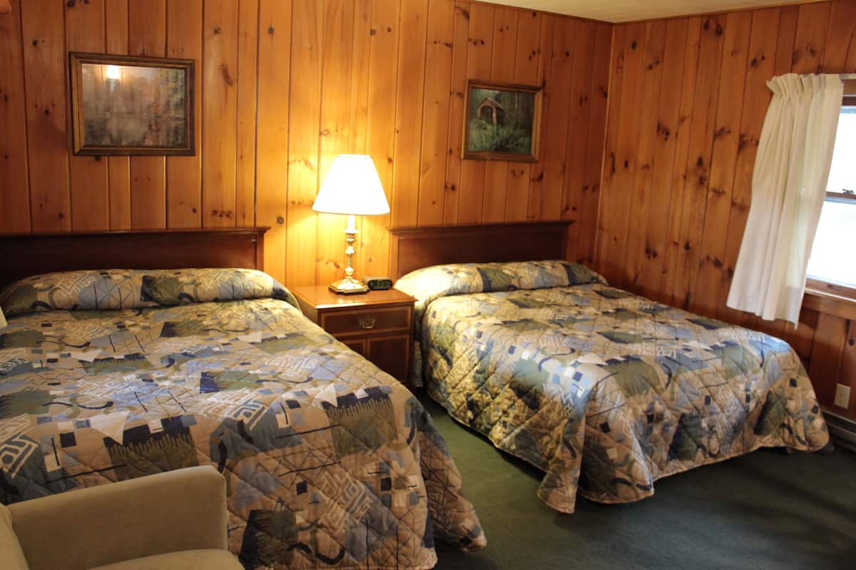 Ein Zimmer mit zwei Betten, Holzwänden, einem Nachttisch mit Lampe und zwei gerahmten Bildern.