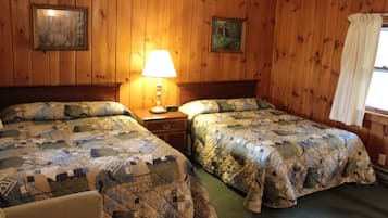 A room with two beds, wooden walls, a nightstand with a lamp, and two framed pictures.