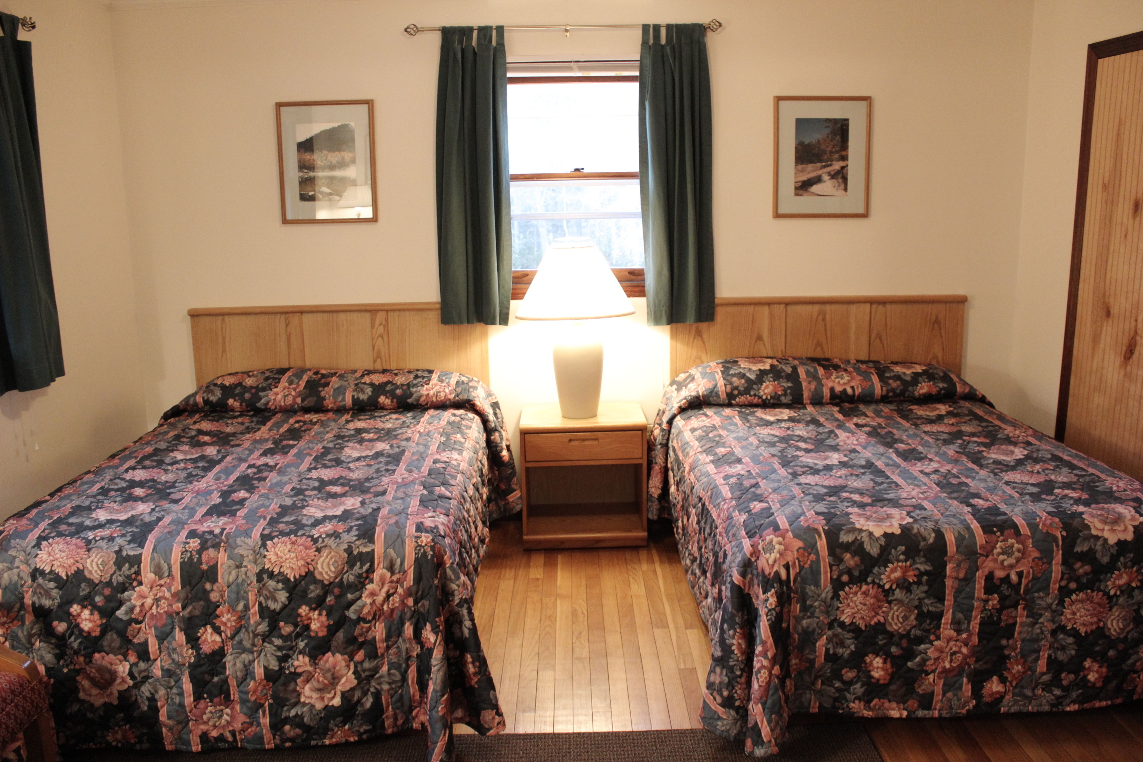 A room with two beds, a wooden headboard, a nightstand with a lamp, and two framed pictures on the walls.