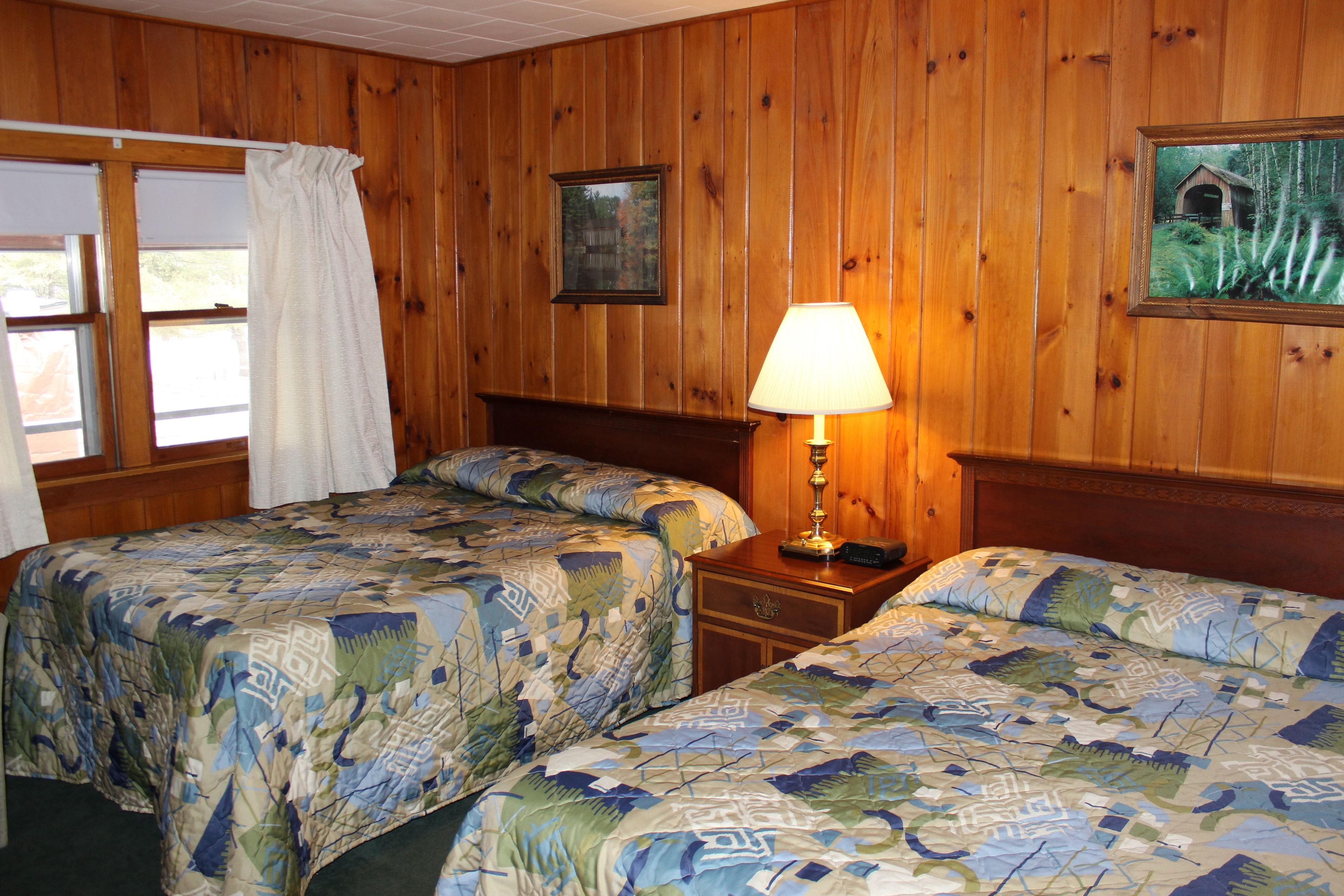 Une chambre avec deux lits, des murs en bois, une fenêtre avec des rideaux, une table de chevet avec une lampe et des tableaux encadrés accrochés au mur.