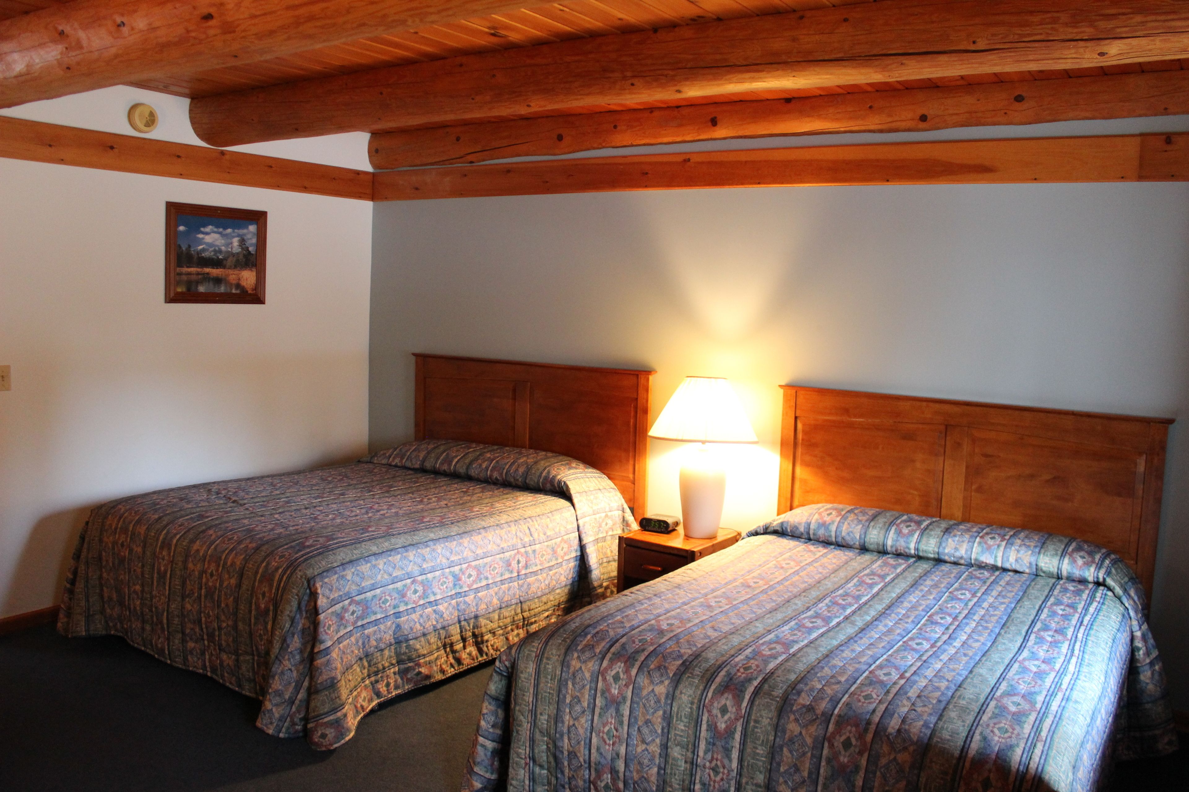A room with two beds, wooden headboards, a nightstand with a lamp, and a framed picture on the wall.