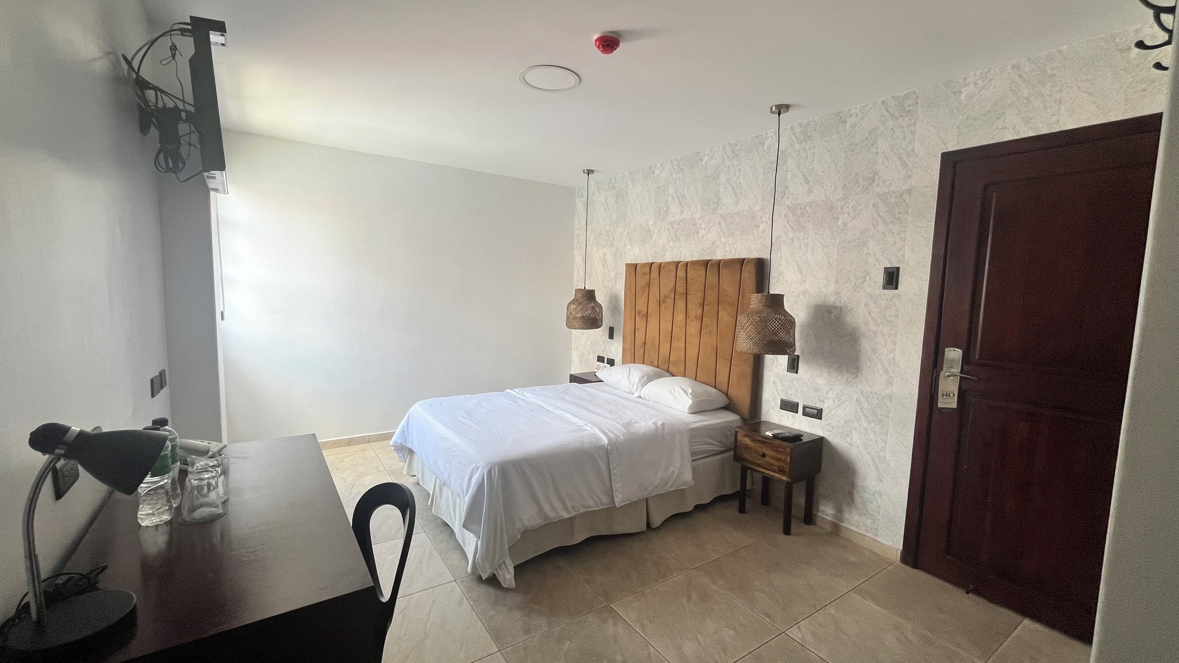 A hotel room with a bed, bedside tables, a desk with a chair, a door, and a wall-mounted light fixture.