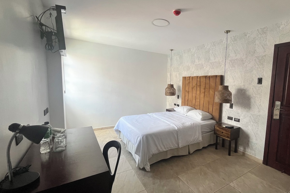 Habitación de hotel con cama, mesitas de noche, un escritorio con silla, una puerta y una lámpara fijada a la pared.