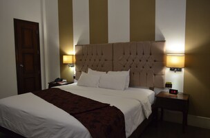 Una habitación de hotel con una cama grande, dos mesitas de noche, una mesita auxiliar, un reloj de pared y una puerta.