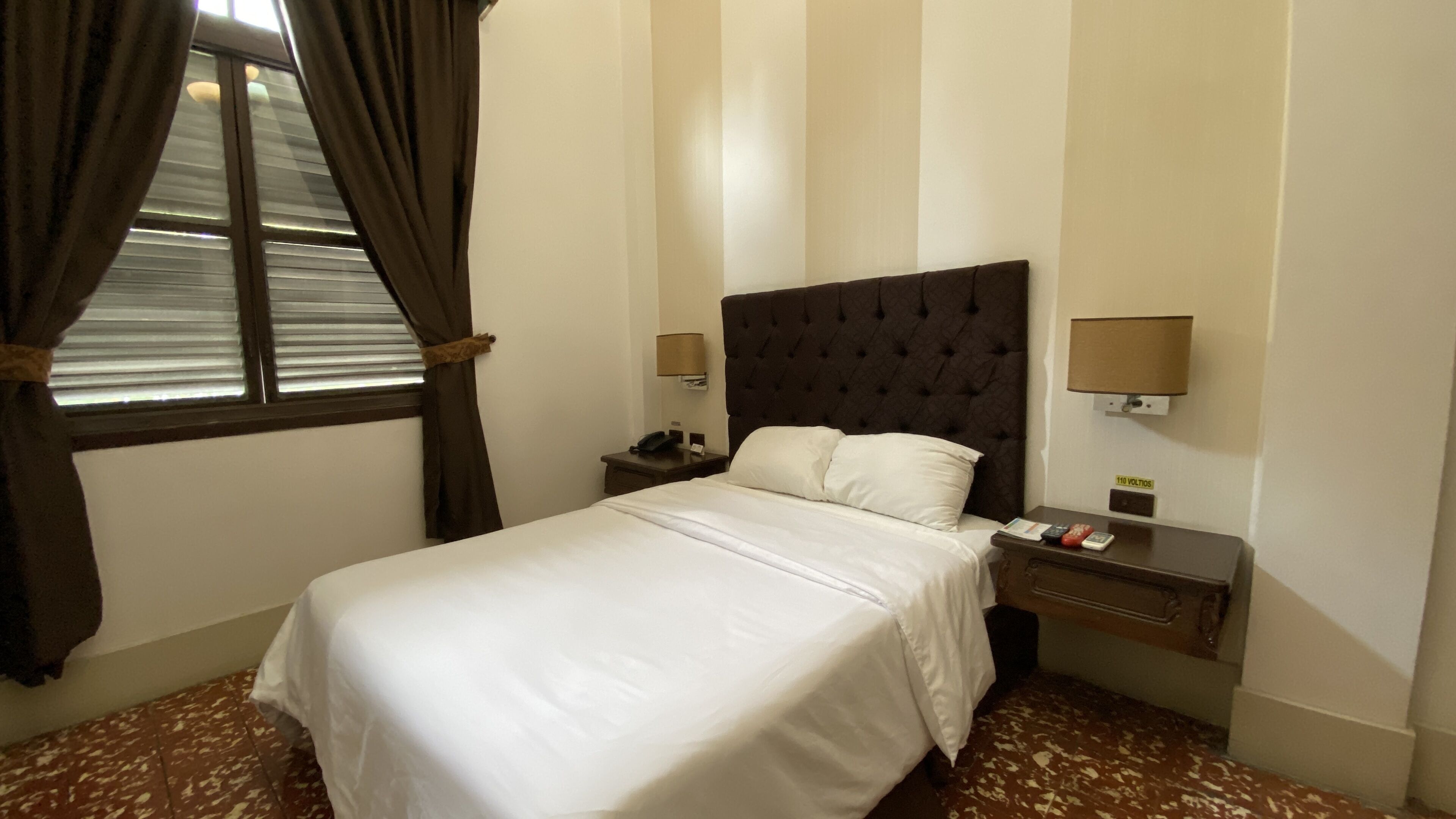 A hotel room with a large bed, two bedside tables, a nightstand, a lamp, and a window with curtains.