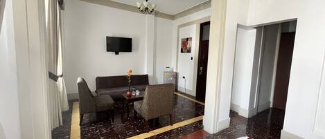 Una habitación de hotel moderna con sofá, dos sillones, una mesita con un florero, televisor de pantalla plana en la pared y alfombra roja.