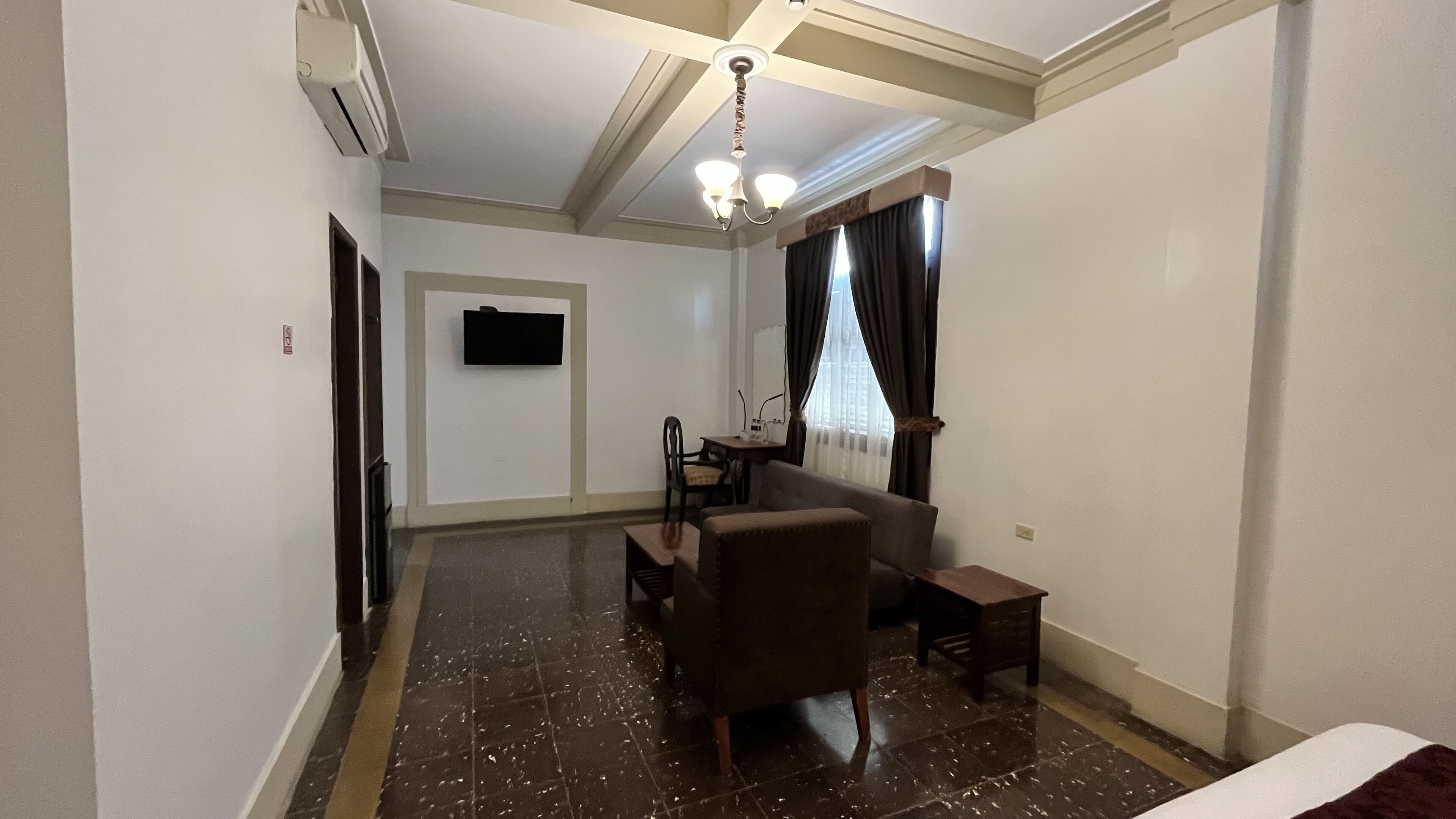 A hotel room with a sofa, a small table, a TV mounted on the wall, a ceiling light fixture, and a window with curtains.