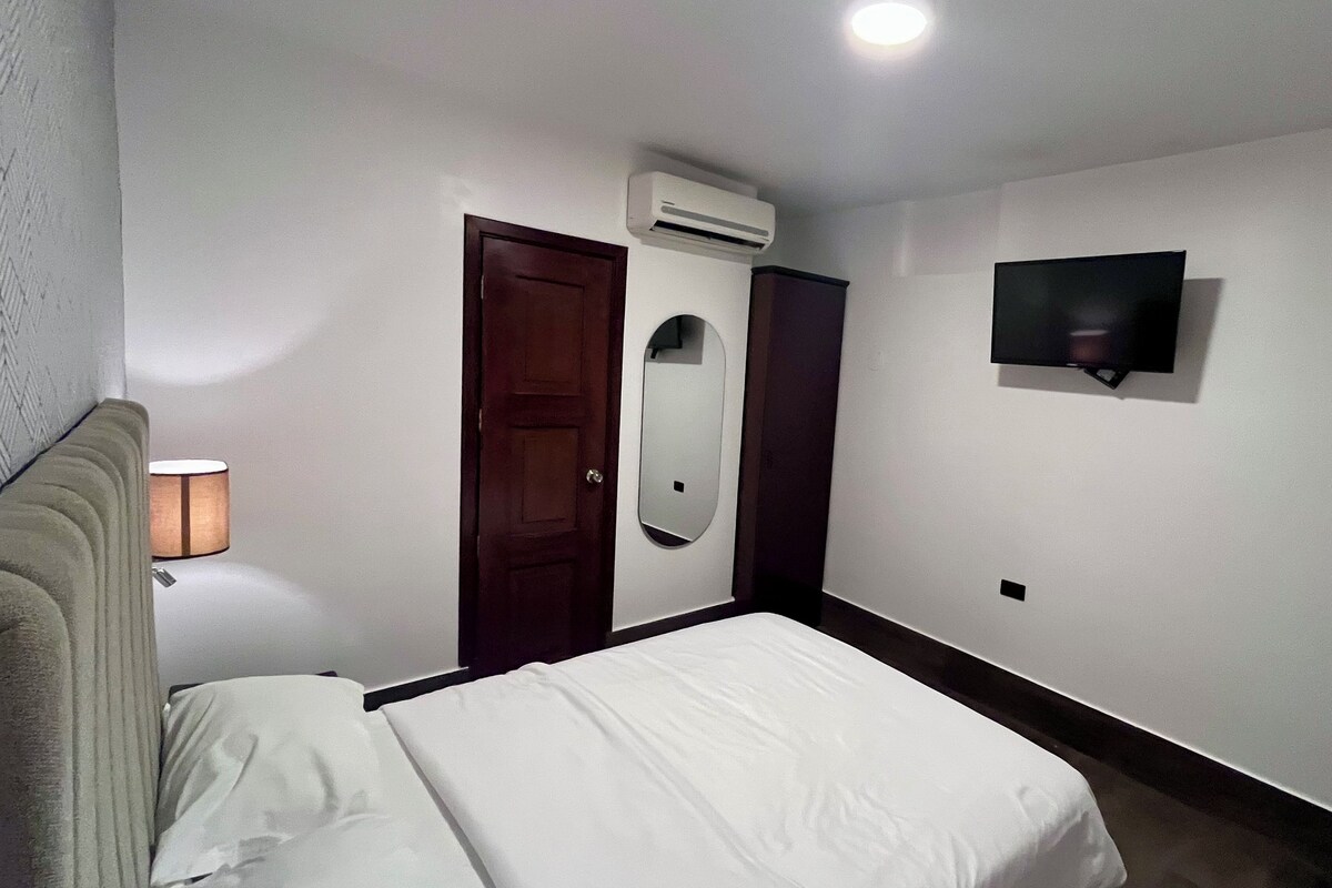 A hotel room with a bed, a wall-mounted TV, an air conditioning unit, a door, and a mirror.
