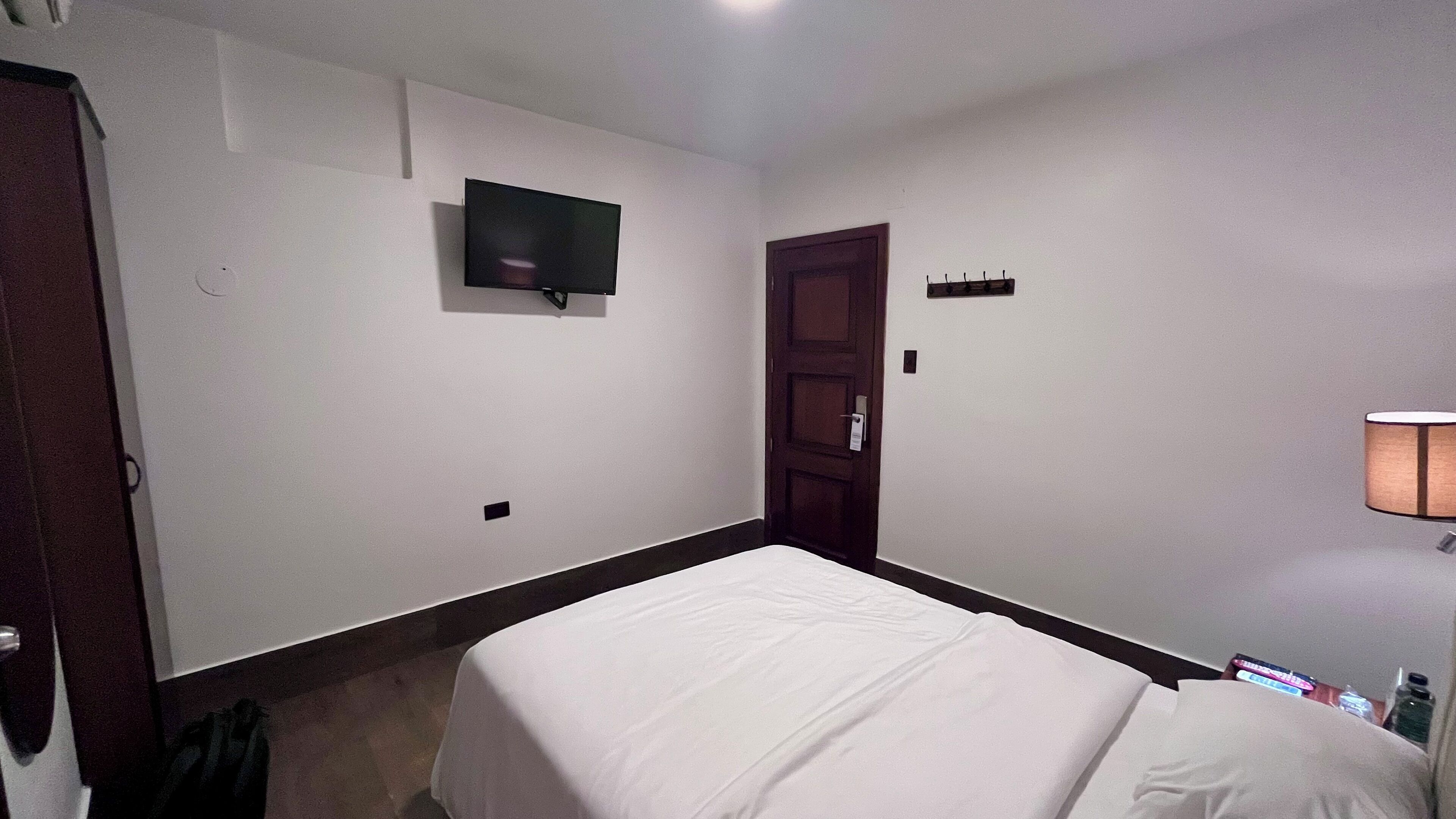 Een hotelkamer met een eenpersoonsbed, een aan de muur bevestigde televisie, een deur en een nachtkastje met een lamp.