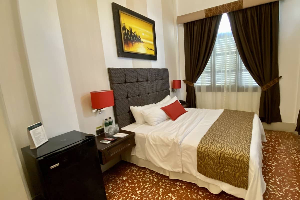 Una habitación de hotel con cama, mesita de noche, escritorio, lámpara, un cuadro y ventana con cortinas.