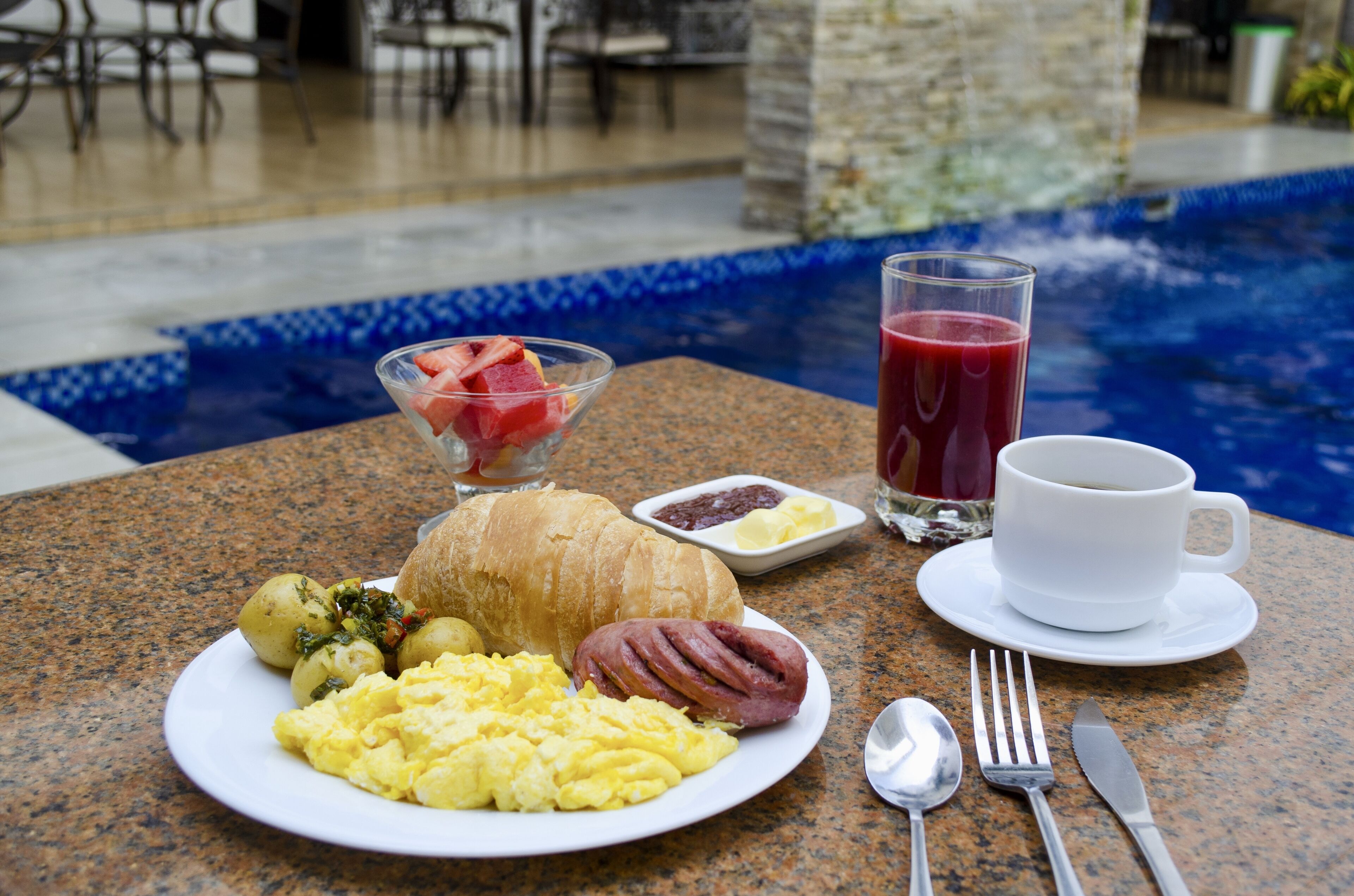A breakfast setting with a croissant, ham, scrambled eggs, and potatoes on a plate, a glass of red juice, and a cup of coffee on a table by a pool.