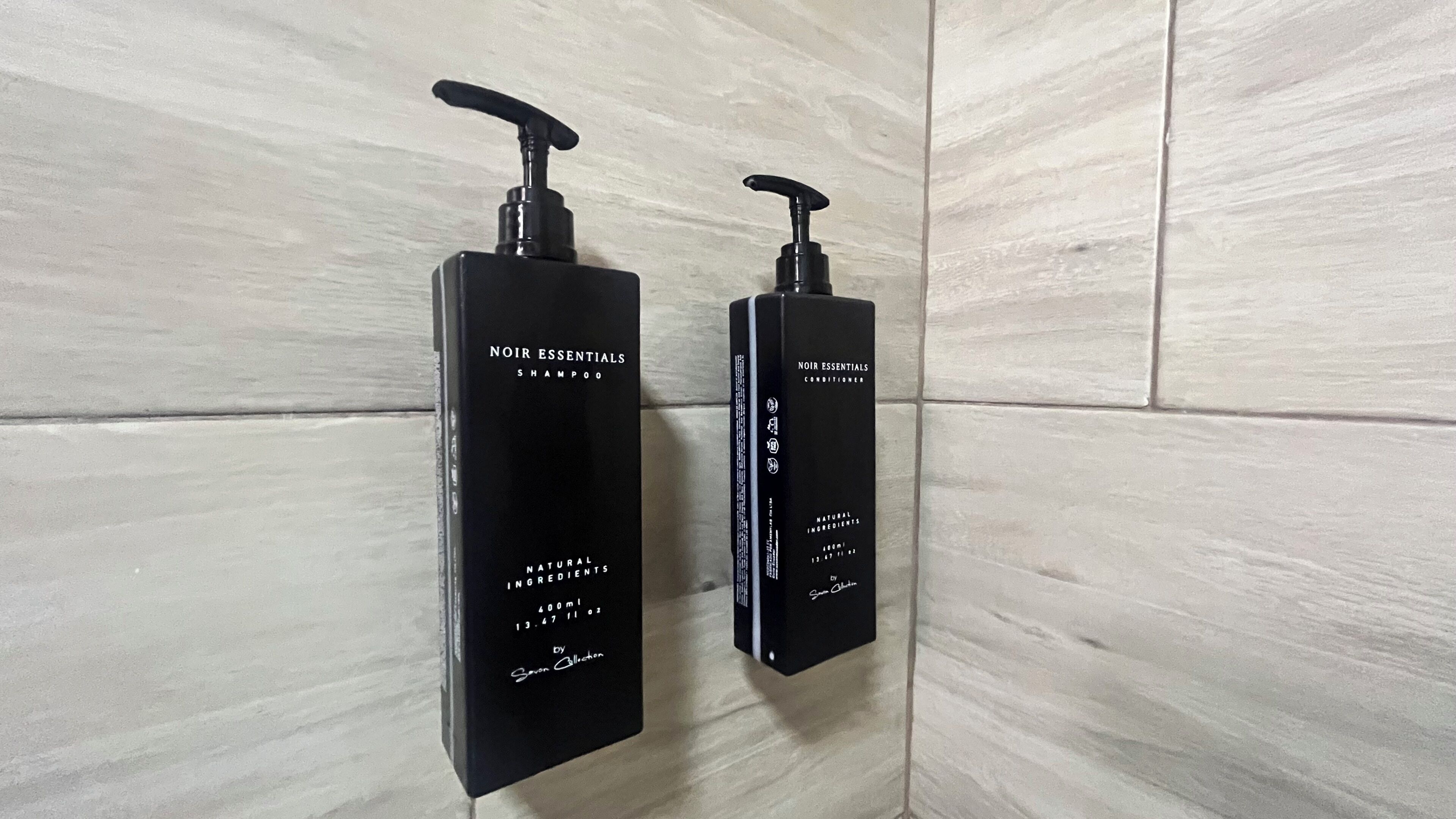 Two black bottles of Noir Essentials Shampoo and Conditioner on a tiled wall.