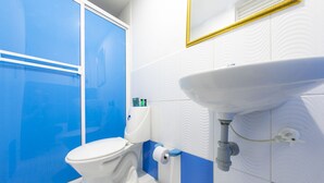 Une salle de bain avec des toilettes blanches, un espace de douche bleu et un lavabo blanc surmonté d’un miroir à cadre doré.