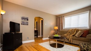 A living room with a television on a black cabinet, a coffee table with apples, a sofa with a leopard print blanket, and a sign on the wall reading "Broadway Wall St." - Perfect Place and Breakfast Too! (Pittsburgh)