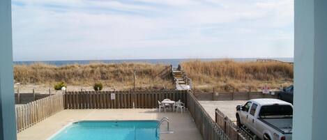 A pool area with a view of the beach, a wooden deck, and a white pickup truck parked nearby.