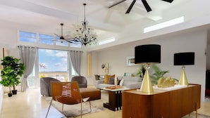 A modern living room with a sofa, armchair, and coffee table. There is a large window with curtains, a ceiling fan, and a chandelier. - Villa Maxine Retreat: Mountainview Pool Near San Juan Airport (Guaynabo)