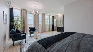 A modern bedroom with a large bed, a sitting area with a chair and a small table, a large window with curtains, and a mirror on the wall. - Villa Maxine Retreat: Mountainview Pool Near San Juan Airport (Guaynabo)