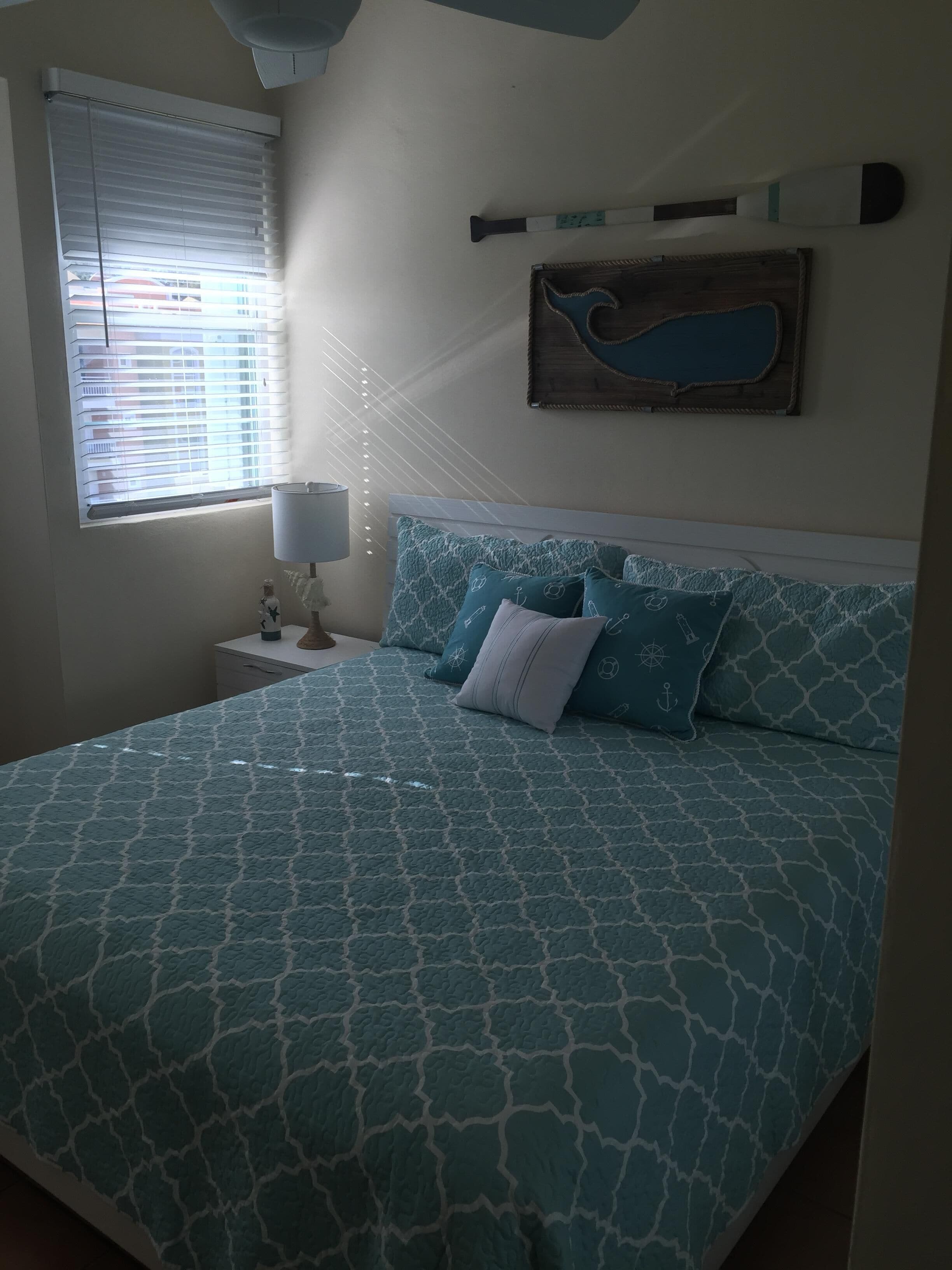Une chambre à coucher comprenant un lit, une table de chevet, une lampe, une fenêtre avec des stores et un élément de décoration fixé au mur.