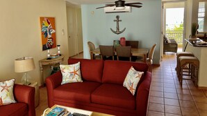 Sala de estar com sofá vermelho, mesa de centro de madeira e ventoinha no teto. Há uma área de jantar com mesa e cadeiras, e uma cozinha com bancada e banquetas.