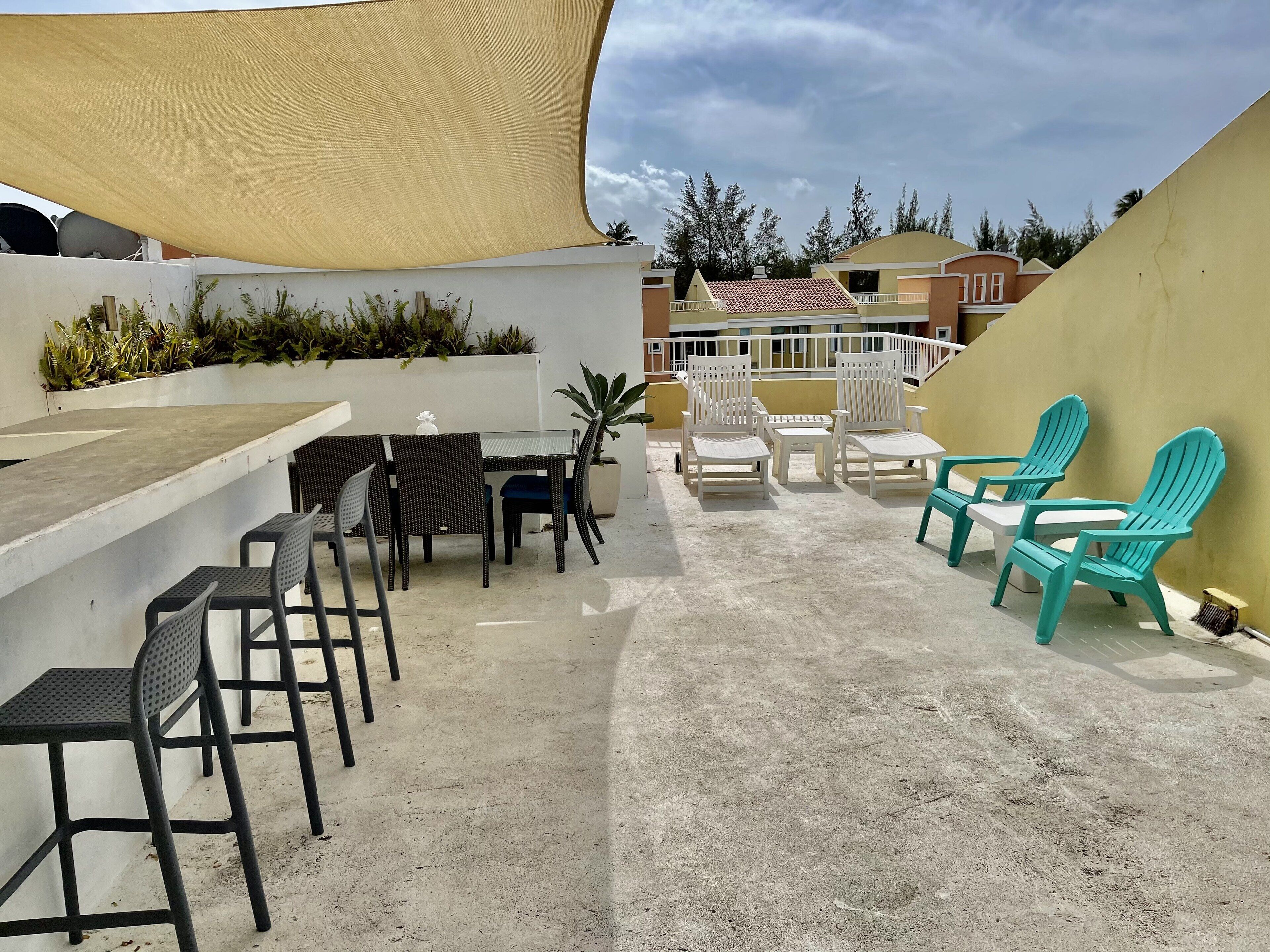 A rooftop terrace with a concrete floor, outdoor seating area with chairs and a table, a large shade sail, and a view of buildings in the background.