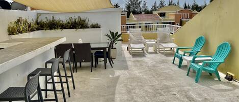 A rooftop terrace with a concrete floor, outdoor seating area with chairs and a table, a large shade sail, and a view of buildings in the background.
