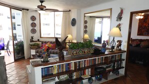 Una sala de estar con suelo de baldosas, un sofá, una mesa de centro con objetos decorativos, una estantería y un ventanal con cortinas.