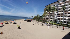 Una playa con un hotel al fondo, palmeras y sombrillas.