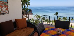 Een balkon met uitzicht op zee, een rieten bank, een tafel met een kleurrijk tafelkleed en een decoratieve schaal.