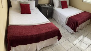 Une chambre d’hôtel avec deux lits, chacun agrémenté de draps blancs et de coussins décoratifs rouges. On y trouve des tables de chevet avec des lampes, et une télécommande posée sur l’une d’elles. La chambre est dotée d’un sol carrelé et de fenêtres équipées de stores.