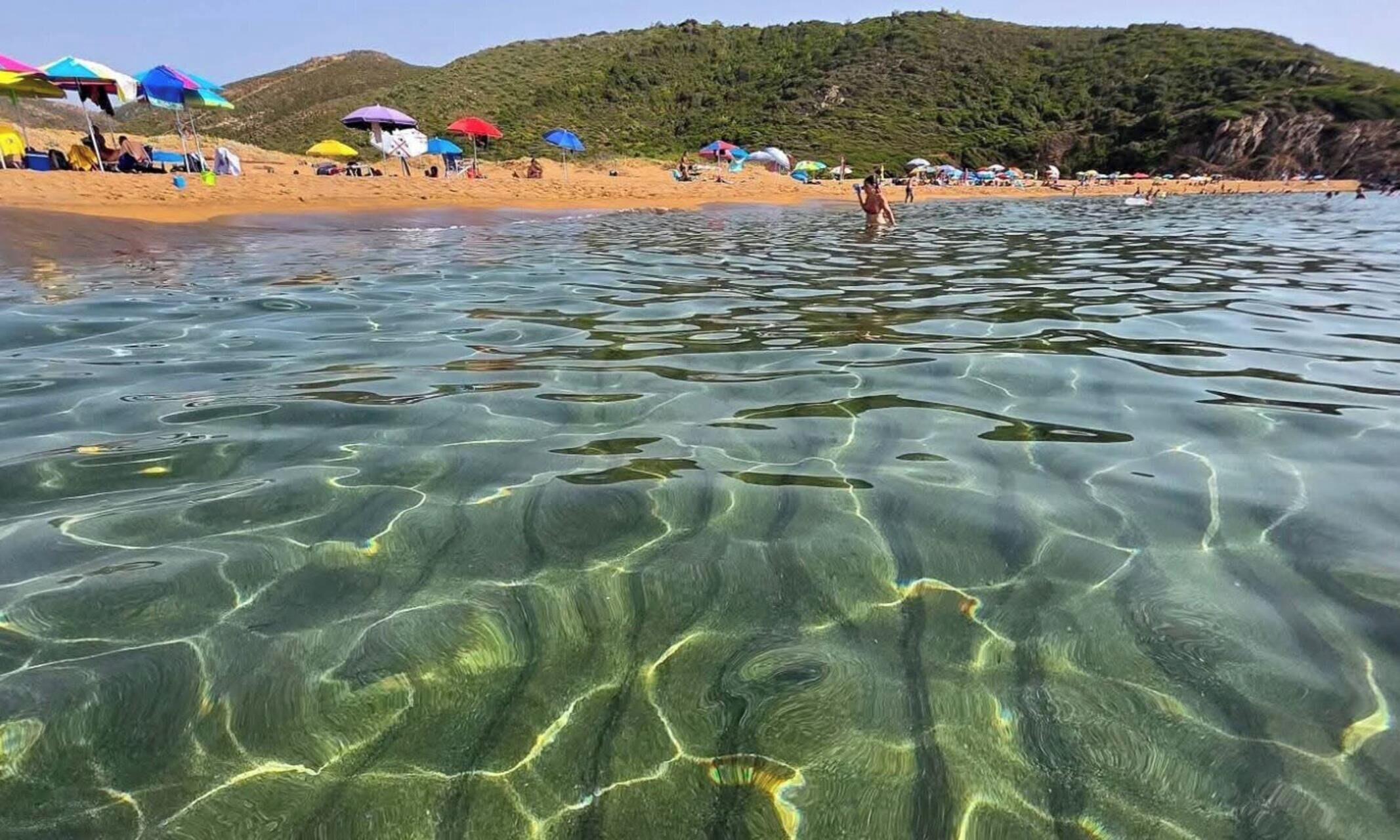 Una spiaggia con acque limpide, ombrelloni e un paesaggio collinare sullo sfondo.