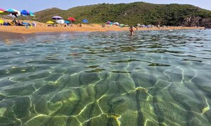 Une plage à l’eau claire, avec des parasols, et un paysage vallonné en arrière-plan.
