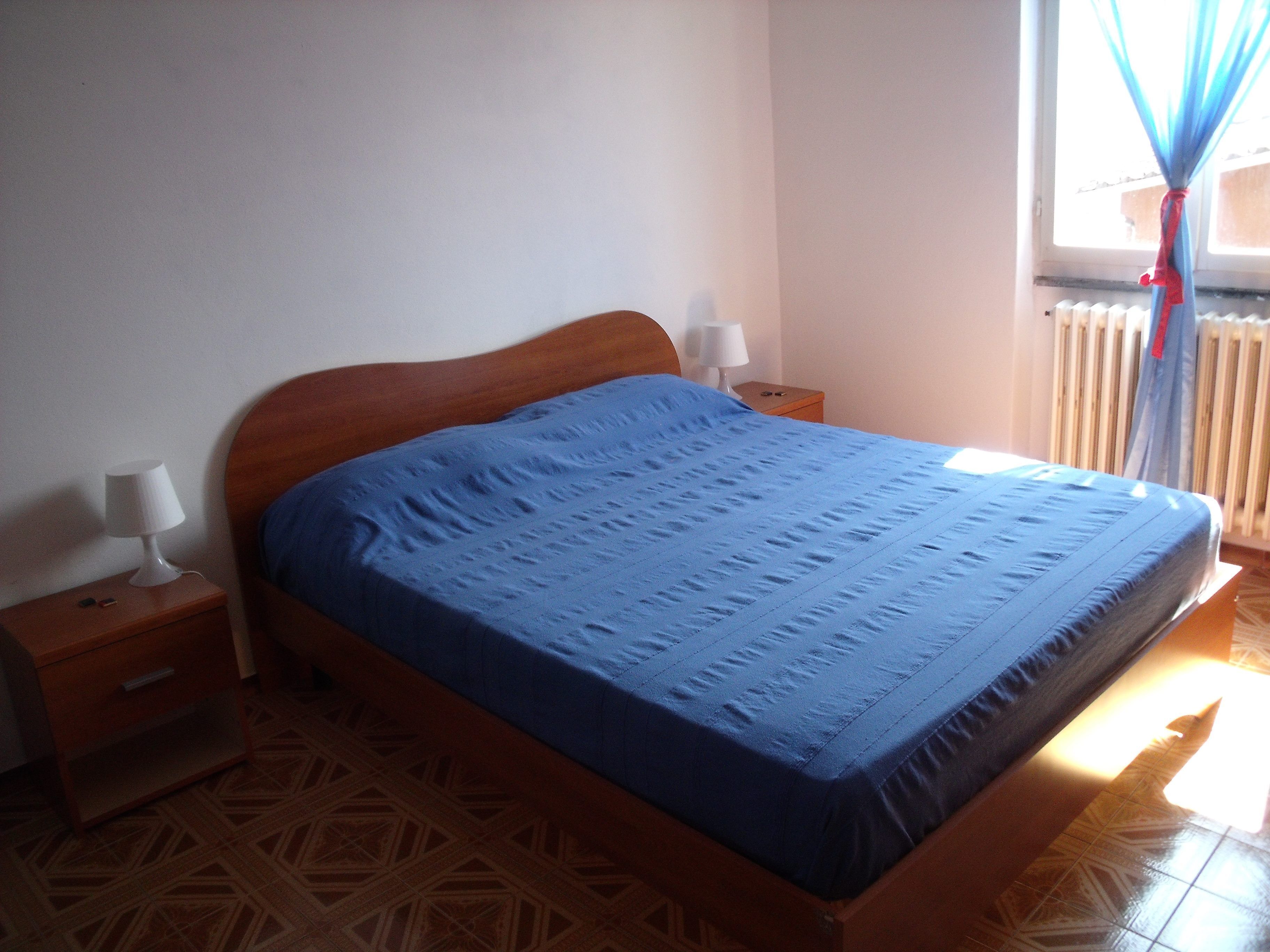 Un dormitorio con una cama de madera, una mesita de noche con lámpara y una ventana con cortinas.