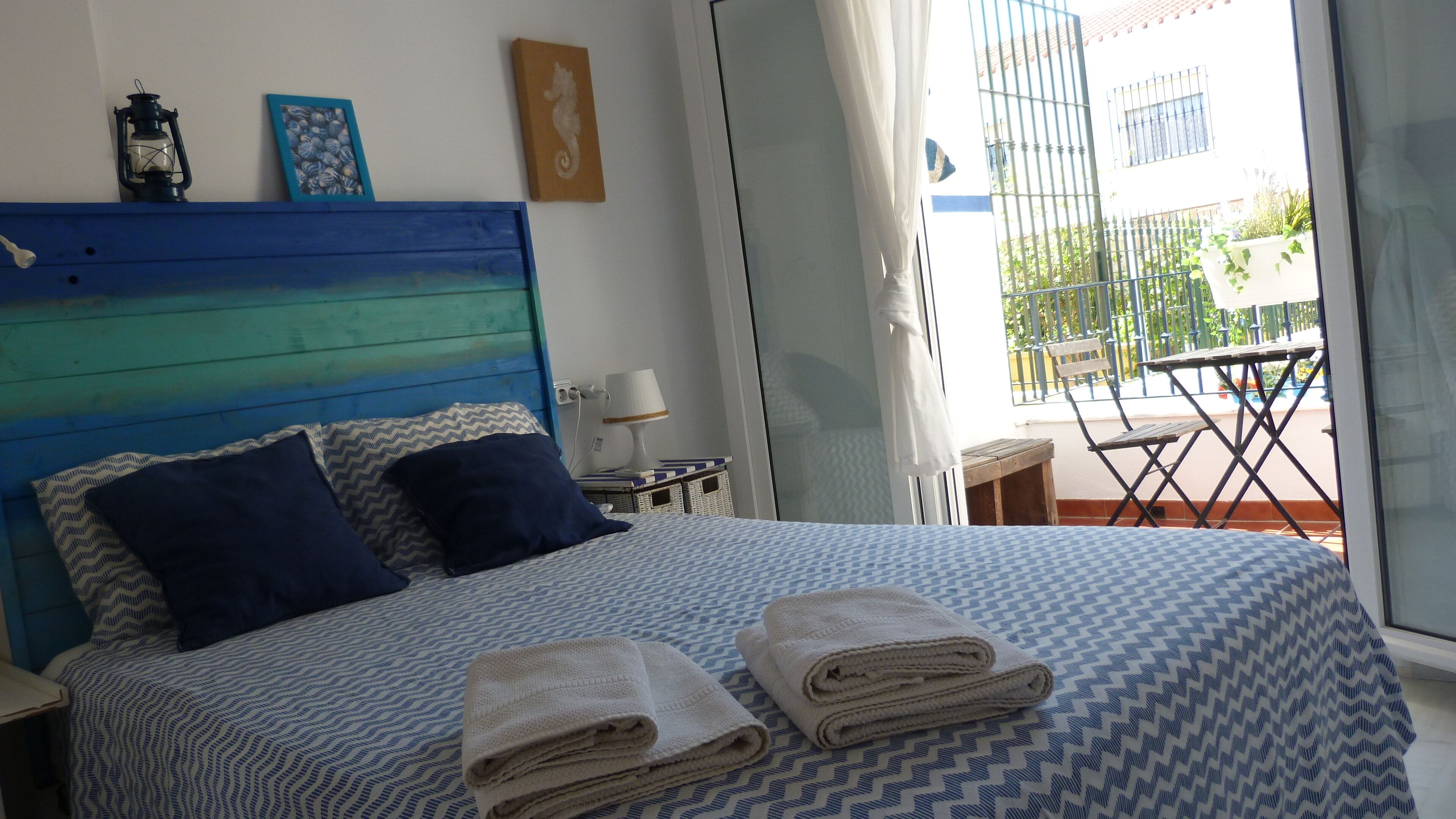 Ein Schlafzimmer mit blauem Kopfteil, zwei Kissen, einem Bett mit gemustertem Überwurf, zwei zusammengerollten Handtüchern, einem Nachttisch mit Lampe und Blick auf den Außenbalkon.