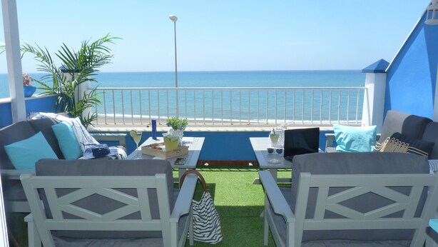 Un balcon avec vue sur la mer, deux chaises blanches, une table avec des boissons et une rambarde bleue.