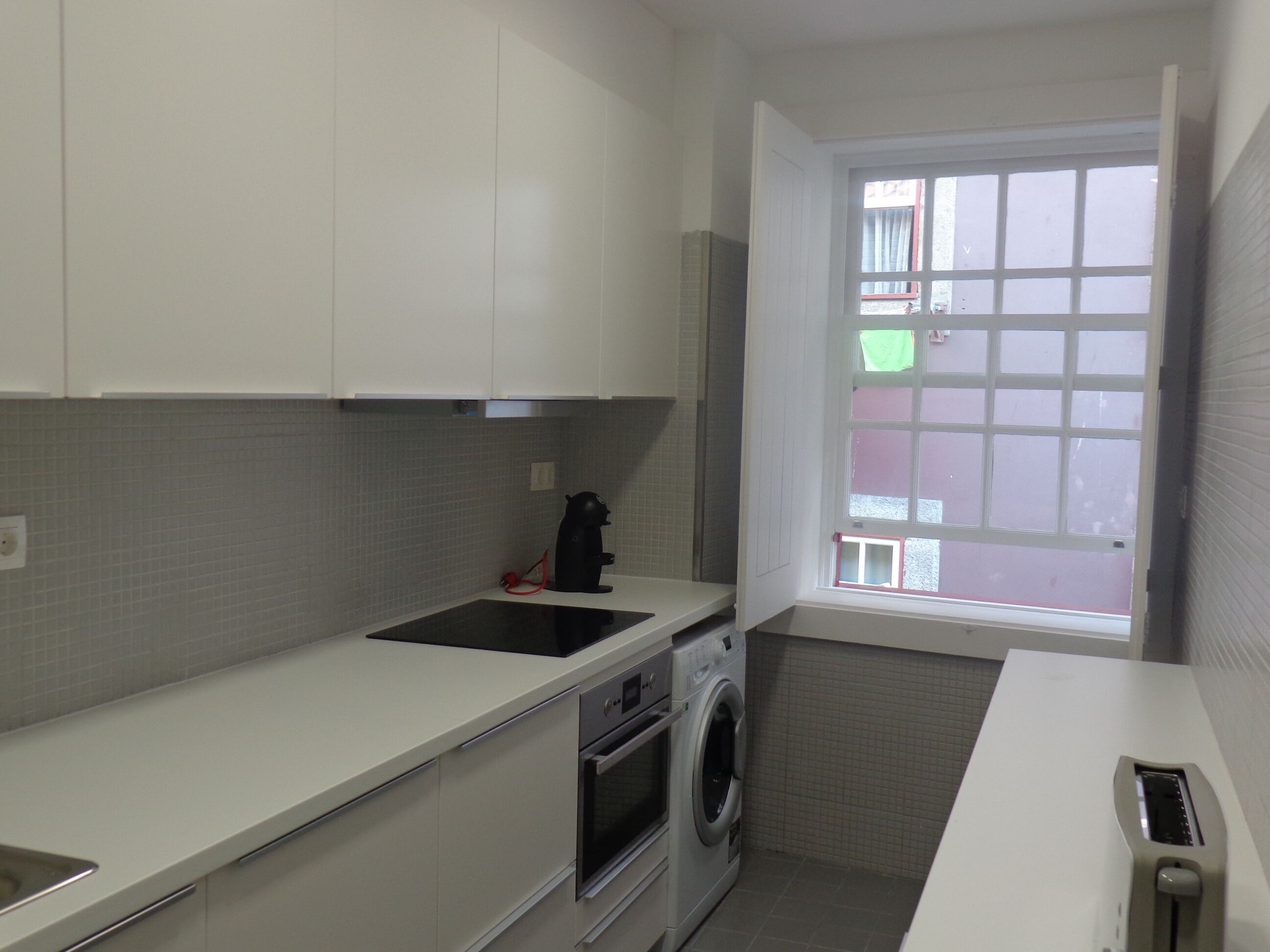 A compact kitchen with white cabinets, a built-in oven, a washing machine, and a window with a view of the street.