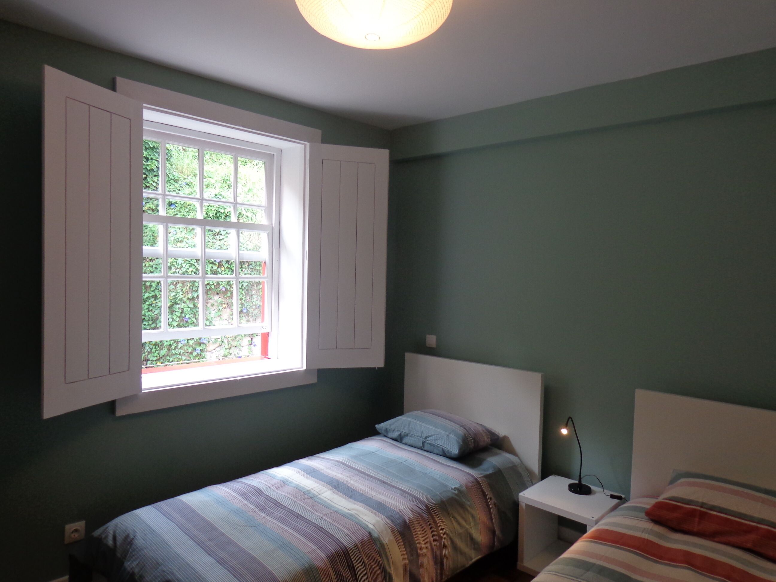 A bedroom with a single bed, a nightstand, a lamp, and a window with shutters.