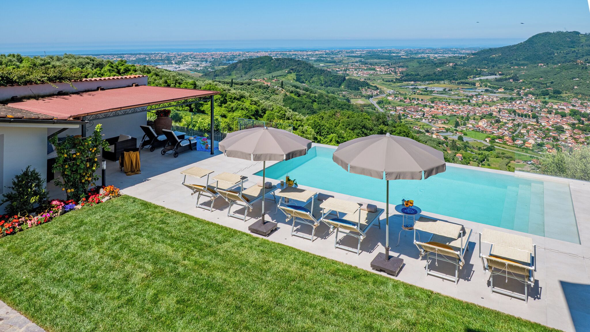 A rooftop pool with lounge chairs and umbrellas, surrounded by a well-maintained lawn and a view of a coastal town.