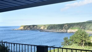 Vista costera desde un balcón techado con barandilla, sobre el mar y los acantilados.