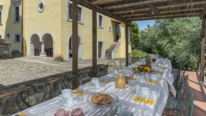 An outdoor dining area with tables set for breakfast, featuring a variety of fruits and pastries, and a building with arches in the background. - Villa Palazzaccio 26, Emma Villas (Arezzo)