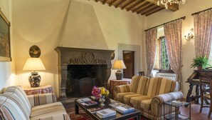 A cozy living room with a fireplace, striped sofas, a coffee table with books, and a red patterned rug. - Villa Palazzaccio 26, Emma Villas (Arezzo)