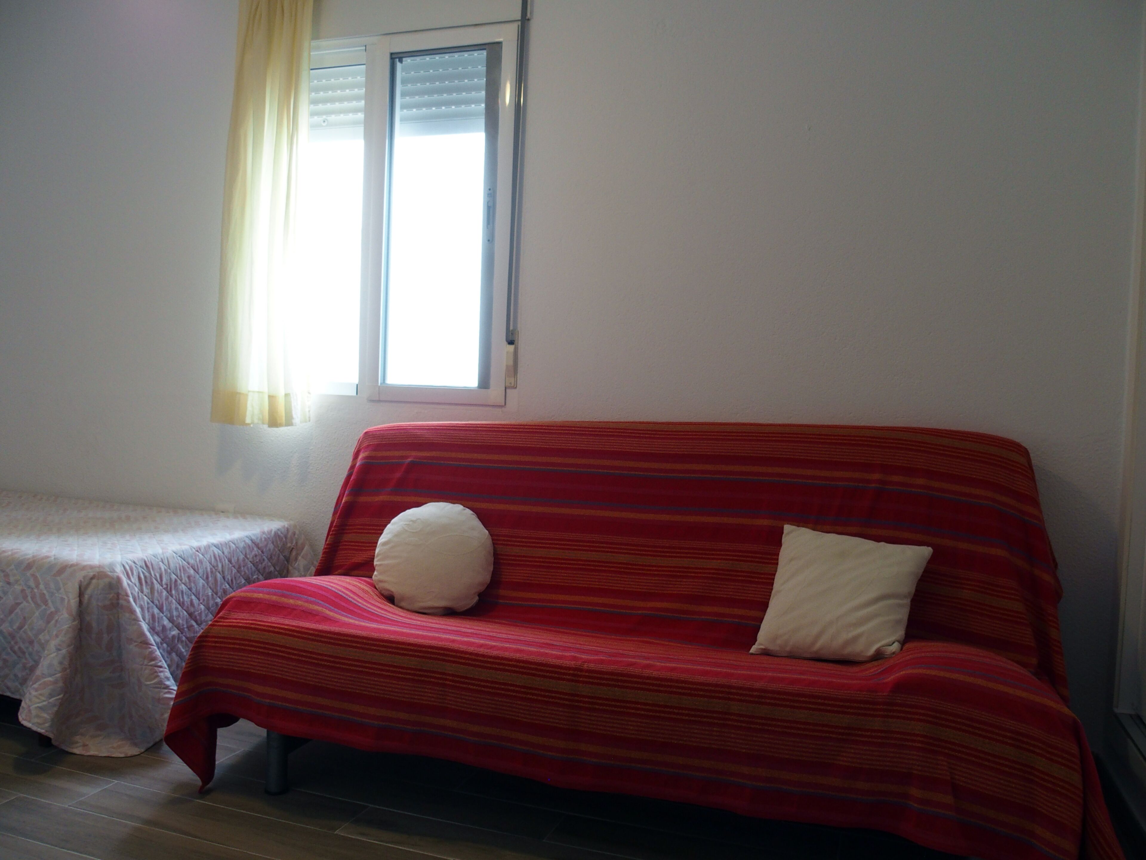 A room with a single bed, a red sofa with two pillows, and a window with blinds.