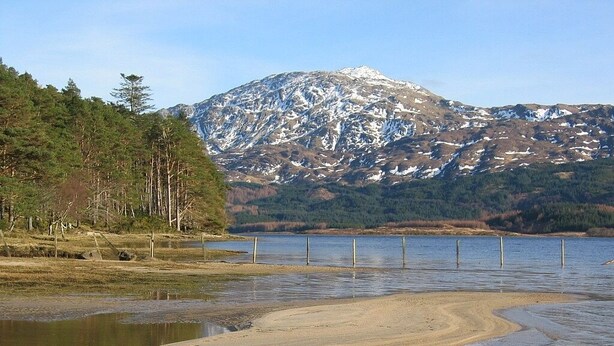 A lake with a sandy shore and a forested area with a mountain in the background. - Traditional Loch Side Croft Cottage And Garden With Contemporary Extension (Acharacle)