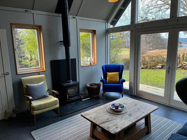 A cozy living room with a fireplace, comfortable seating, and large windows offering a view of the outdoors.