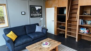 A modern living room with a dark blue sofa, a wooden coffee table, a bookshelf, and a framed travel poster on the wall. - Traditional Loch Side Croft Cottage And Garden With Contemporary Extension (Acharacle)
