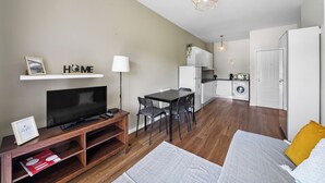 A modern apartment with a flat-screen TV on a wooden stand, a dining area with a table and chairs, a kitchen with a washing machine, and a bed with a yellow pillow. - Apartamento D'alegria 1 by Amber Star Rent (Porto)