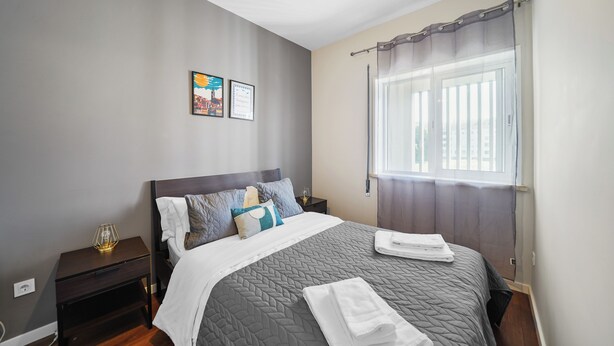 A neatly made bed with a grey quilt, a bedside table with a lamp, and a window with curtains. - Apartamento D'alegria 1 by Amber Star Rent (Porto)