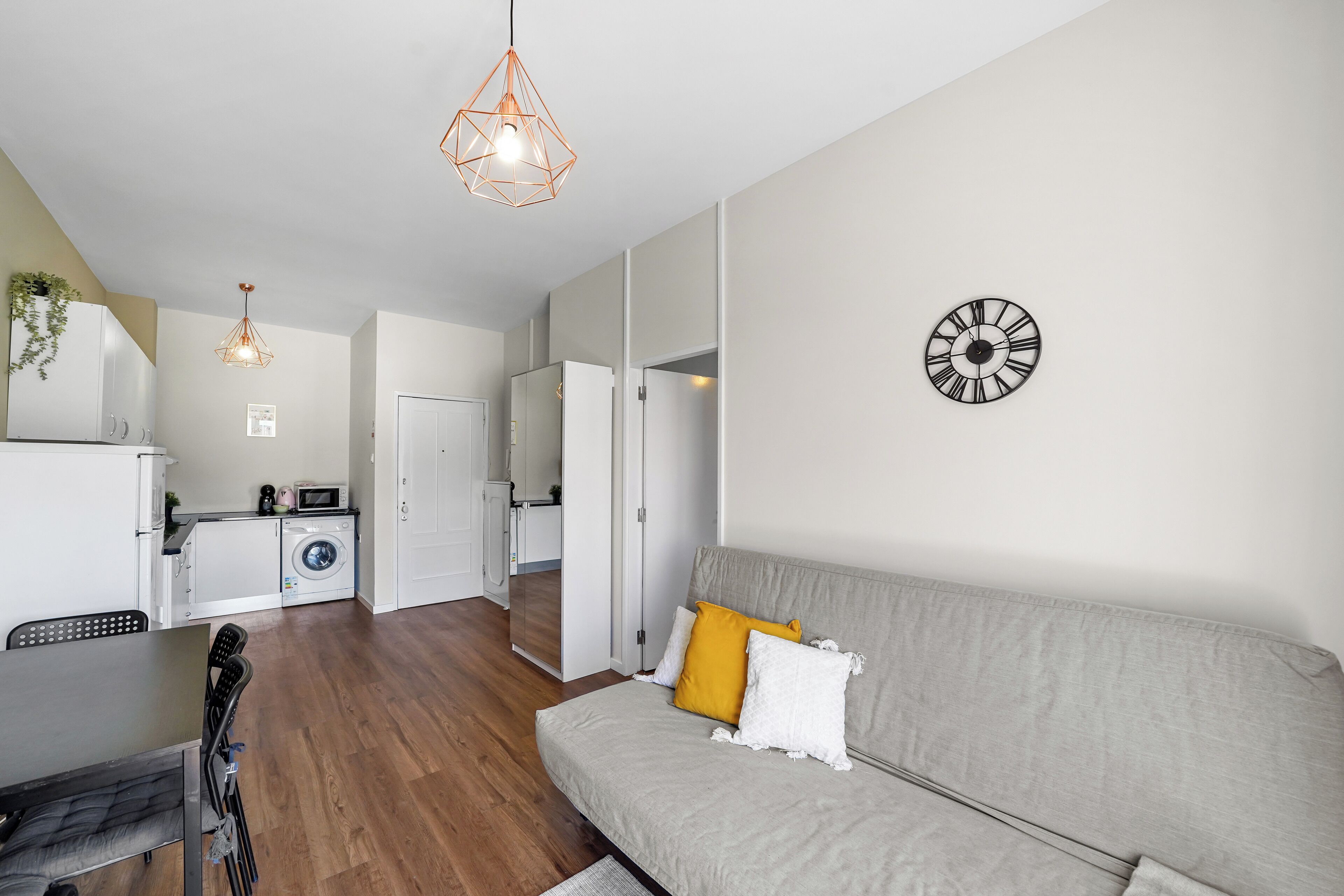 Un salon moderne avec un canapé gris, un coin repas avec une table et des chaises, une cuisine avec des appareils électroménagers blancs et un luminaire suspendu.